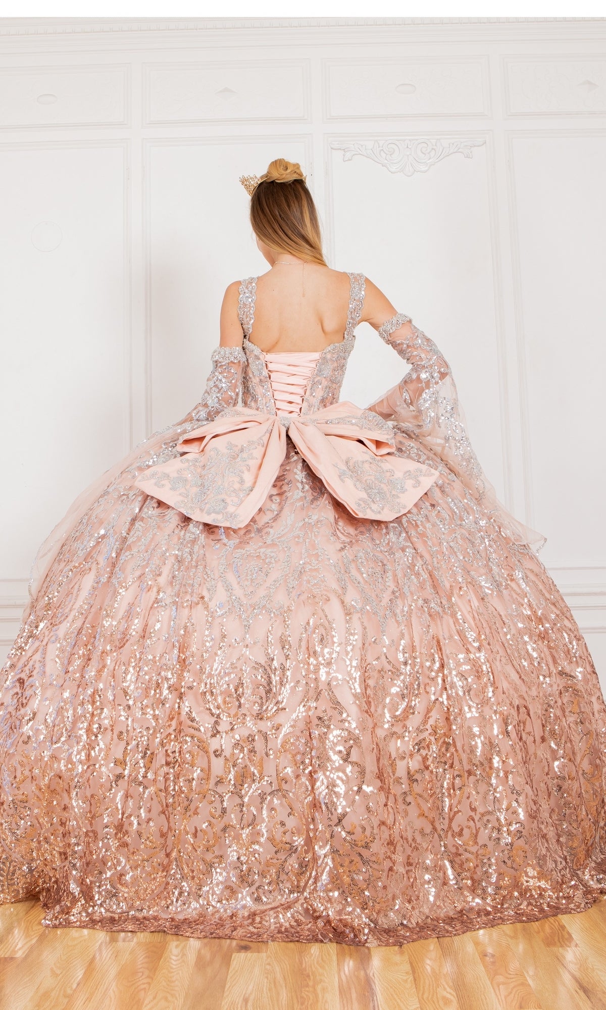 A woman in the Quinceanera Ball Gown 8080J—sparkling pink ombre with silver details and a large bow—faces a white ornate wall, crowned in gold.